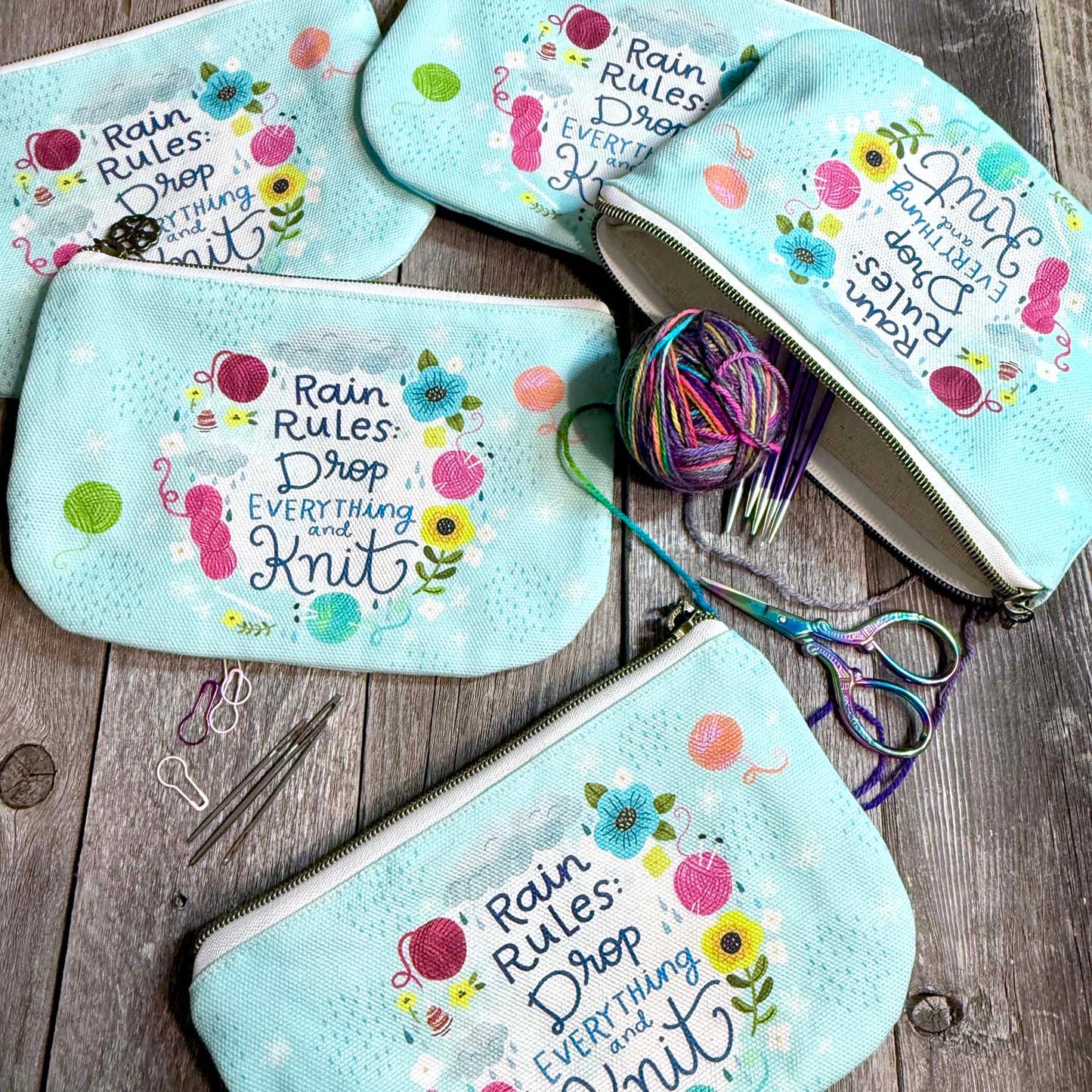 blue green canvas zippered pouch printed with yarn and flowers that says "rain rules drop everything and knit"