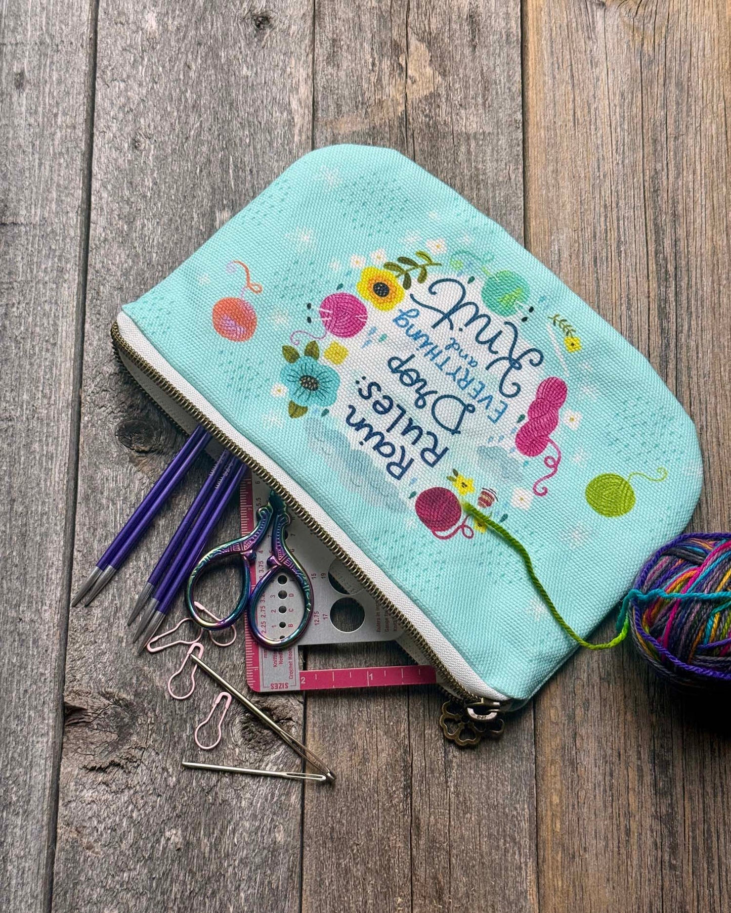 blue green canvas zippered pouch printed with yarn and flowers that says "rain rules drop everything and knit"