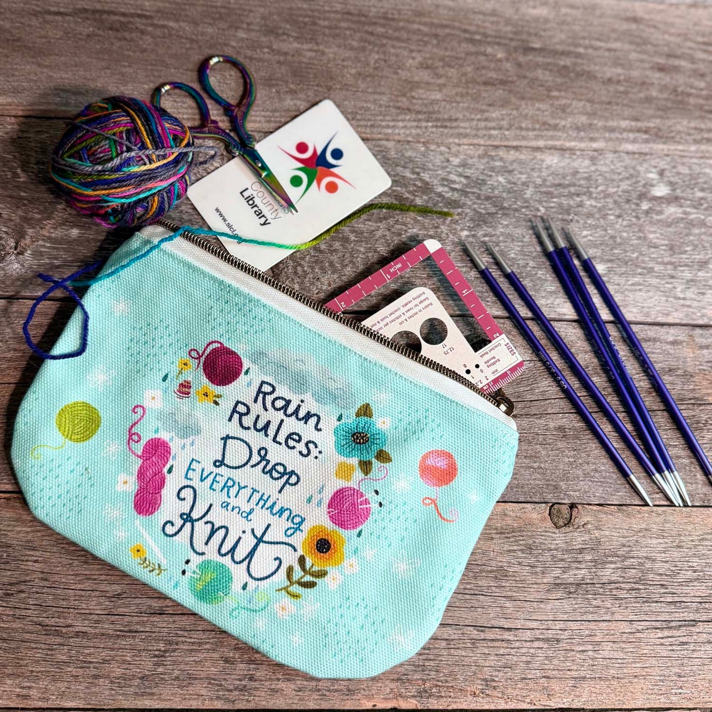 blue green canvas zippered pouch printed with yarn and flowers that says "rain rules drop everything and knit" 