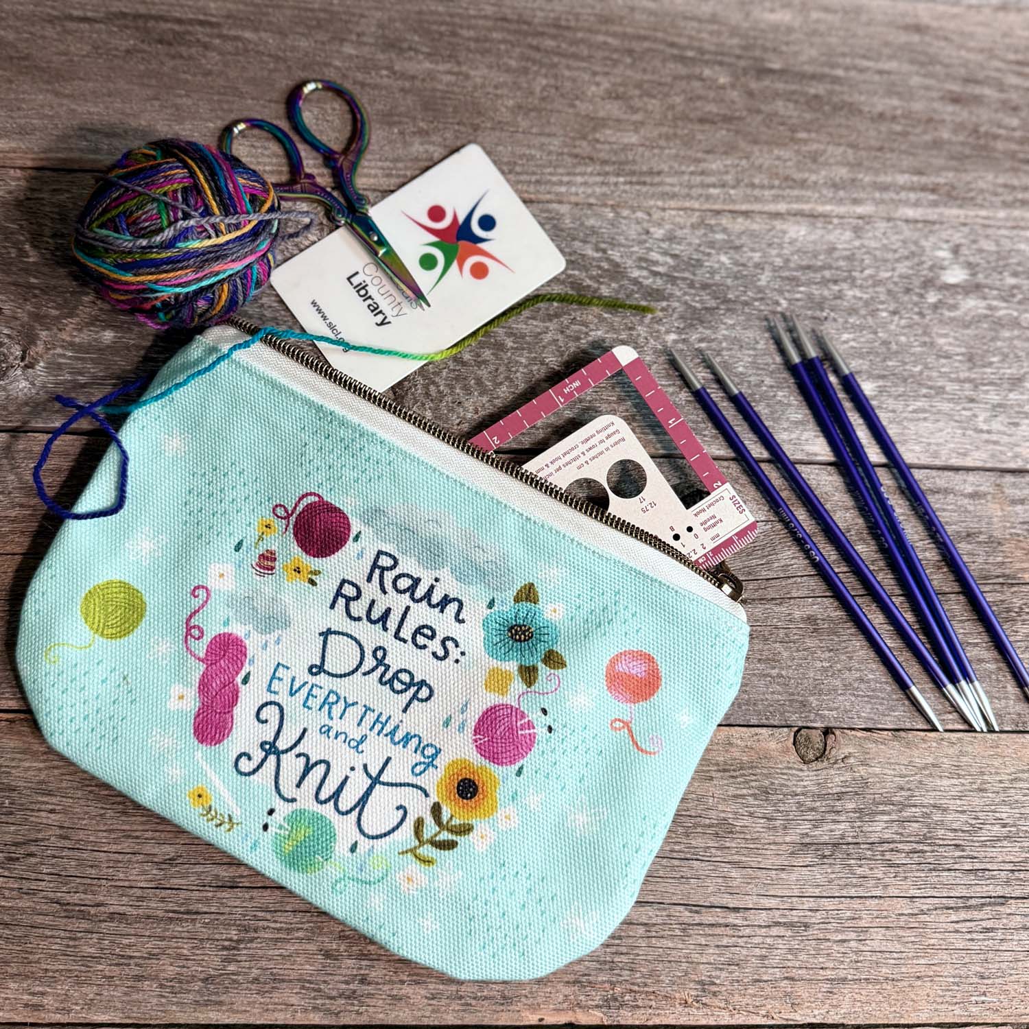 blue green canvas zippered pouch printed with yarn and flowers that says "rain rules drop everything and knit" 