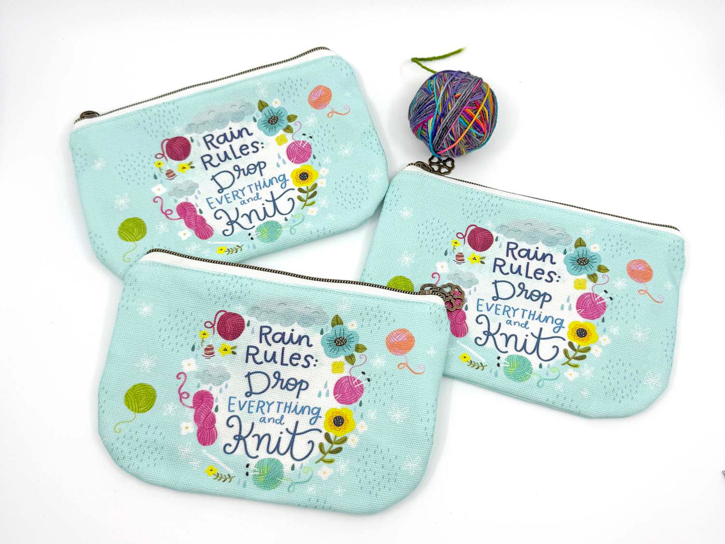 blue green canvas zippered pouch printed with yarn and flowers that says "rain rules drop everything and knit" 